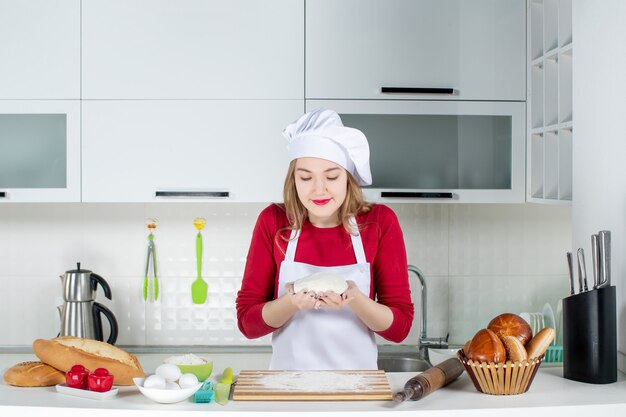 Vista frontal joven cocinera con sombrero de cocinero y delantal sosteniendo la masa con ambas manos en la cocina