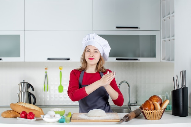 Vista frontal joven cocinera con sombrero de cocinero y delantal uniendo sus manos en la cocina