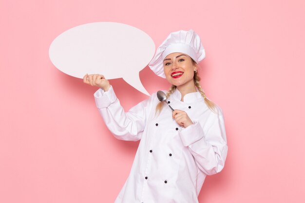 Vista frontal joven cocinera en traje de cocinero blanco con cartel enorme blanco con cuchara en el cocinero espacial rosa