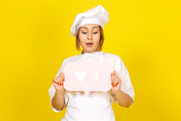 Una vista frontal joven cocinera en traje de cocinero blanco y gorra blanca con cartel rosa en el amarillo