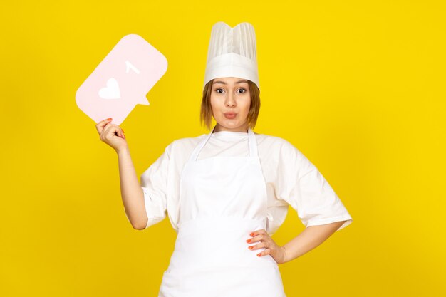 Una vista frontal joven cocinera en traje de cocinero blanco y gorra blanca con cartel rosa posando en el amarillo