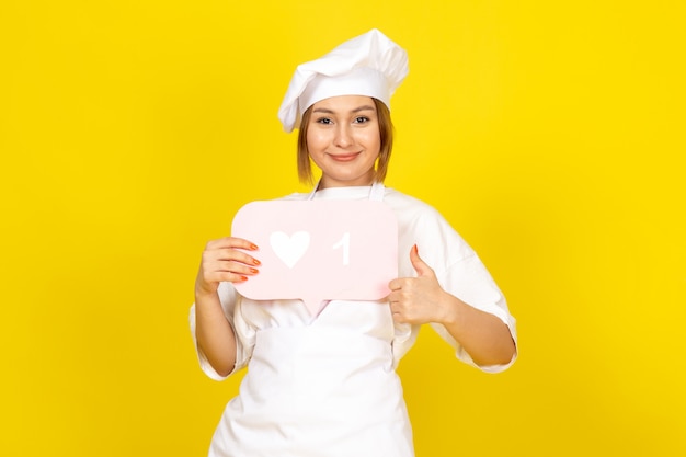 Una vista frontal joven cocinera en traje de cocinero blanco y gorra blanca con cartel rosa sonriendo en el amarillo