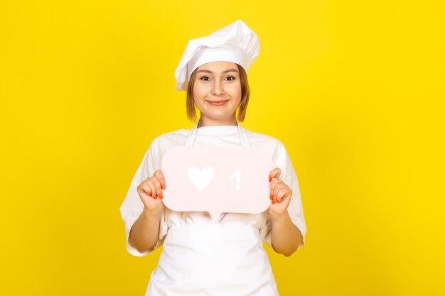 Una vista frontal joven cocinera en traje de cocinero blanco y gorra blanca con cartel rosa sonriendo en el amarillo