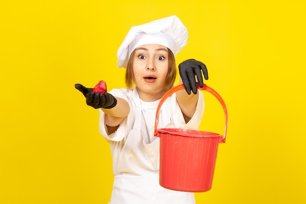 Una vista frontal joven cocinera en traje de cocinero blanco y gorra blanca en guantes negros con cesta roja y fresa en el amarillo