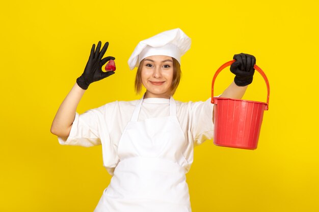 Una vista frontal joven cocinera en traje de cocinero blanco y gorra blanca en guantes negros con cesta roja y fresa sonriendo en el amarillo