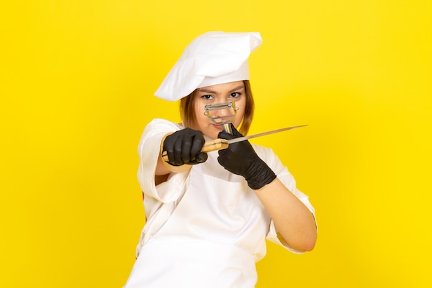 Una vista frontal joven cocinera en traje de cocinero blanco y gorra blanca en guantes negros con cuchillo y limpiador de verduras en el amarillo