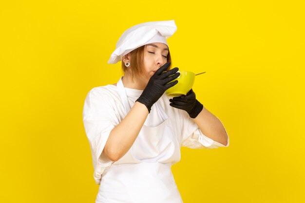 Una vista frontal joven cocinera en traje de cocinero blanco y gorra blanca en guantes negros con placa verde comiendo espagueti en el amarillo