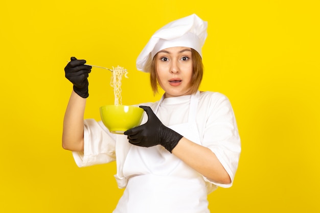 Una vista frontal joven cocinera en traje de cocinero blanco y gorra blanca en guantes negros con placa verde con espagueti en el amarillo