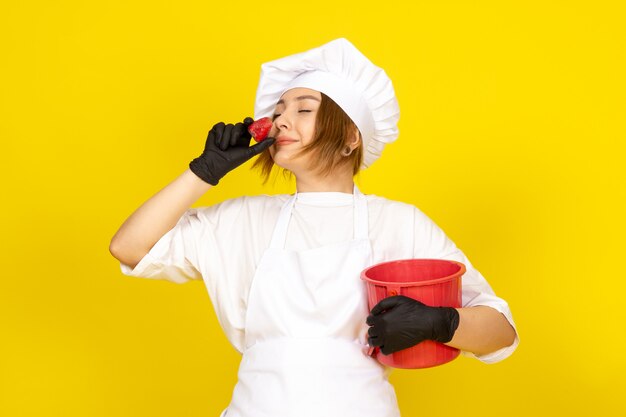 Una vista frontal joven cocinera en traje de cocinero blanco y gorra blanca en guantes negros sosteniendo una canasta roja sonriendo con olor a fresa en el amarillo