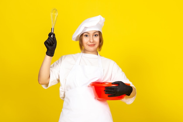 Una vista frontal joven cocinera en traje de cocinero blanco y gorra blanca en guantes negros sosteniendo tazón rojo mezclándolo sobre el amarillo