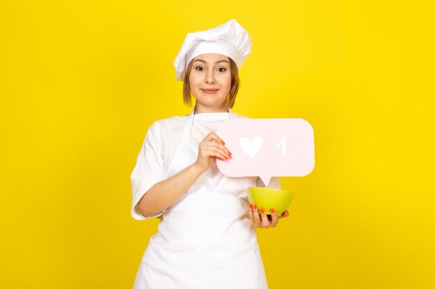 Una vista frontal joven cocinera en traje de cocinero blanco y gorra blanca con placa verde y cartel rosa sonriendo en el amarillo