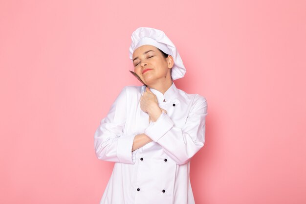 Una vista frontal joven cocinera en traje de cocinero blanco gorra blanca posando sosteniendo una cuchara de plata grande sonriendo expresión encantada