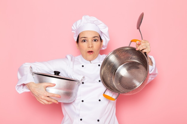Foto gratuita una vista frontal joven cocinera en traje de cocinero blanco gorra blanca posando sosteniendo una gran cuchara de plata y una cacerola de plata expresión sorprendida
