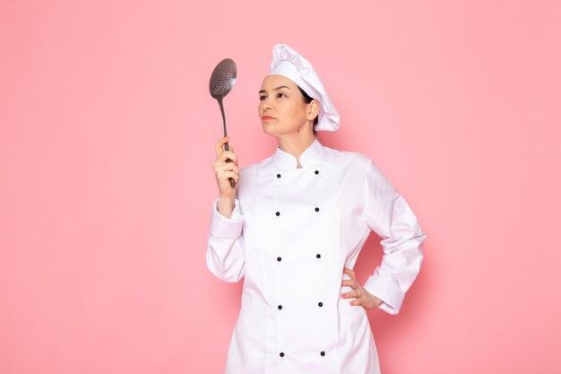 Una vista frontal joven cocinera en traje de cocinero blanco gorra blanca posando sosteniendo una gran cuchara de plata enojado preparándose para golpear