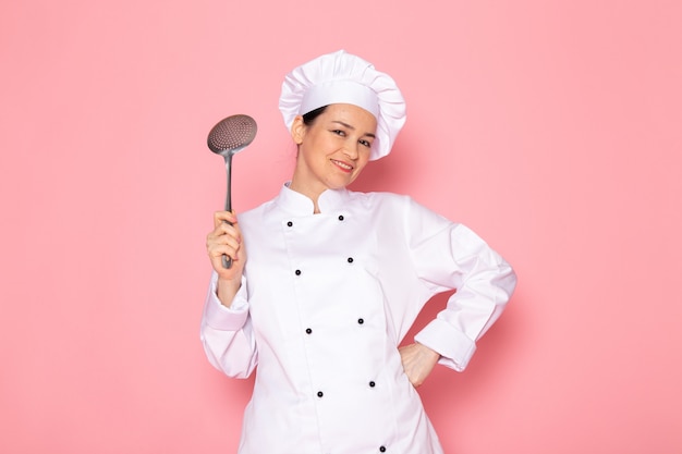 Una vista frontal joven cocinera en traje de cocinero blanco gorra blanca posando sosteniendo gran cuchara de plata sonriendo