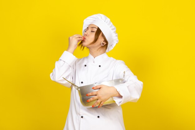 Una vista frontal joven cocinera en traje de cocinero blanco y gorra blanca sosteniendo una bandeja de plata redonda expresando sabroso cartel en el amarillo