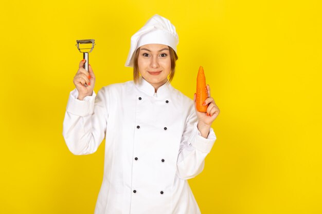 Una vista frontal joven cocinera en traje de cocinero blanco y gorra blanca sosteniendo y limpiando zanahoria naranja sonriendo en el amarillo