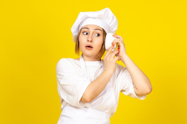 Una vista frontal joven cocinera en traje de cocinero blanco y gorra blanca sosteniendo la taza blanca escuchando la copa en el amarillo
