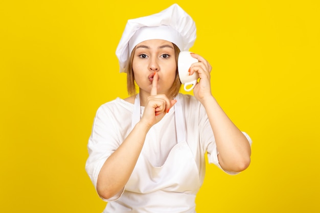 Una vista frontal joven cocinera en traje de cocinero blanco y gorra blanca sosteniendo una taza blanca escuchando la taza mostrando señal de silencio en el amarillo