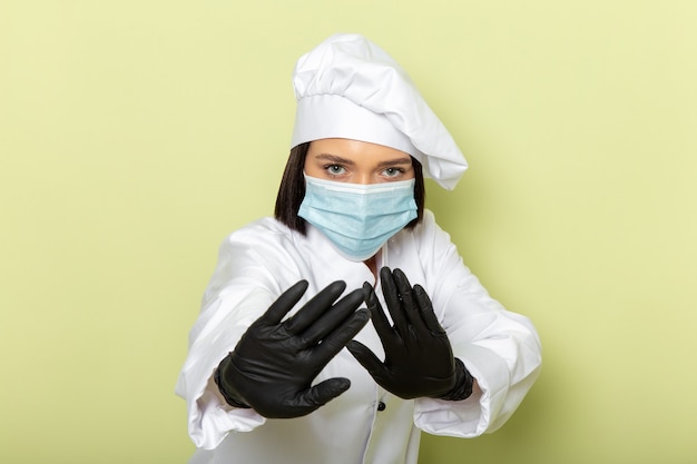 Una vista frontal joven cocinera en traje de cocinero blanco y gorra con guantes y máscara estéril con pose cautelosa en la pared verde dama trabajo color de alimentos