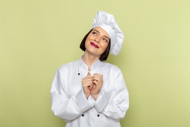 Vista frontal de una joven cocinera en traje de cocinero blanco y gorra posando y sonriendo en la pared verde dama trabajo cocina color