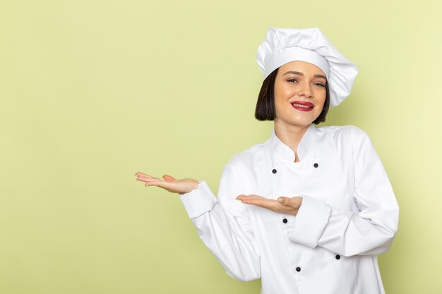 Una vista frontal joven cocinera en traje de cocinero blanco y gorra posando con una sonrisa en la pared verde dama trabajo color de la cocina de alimentos