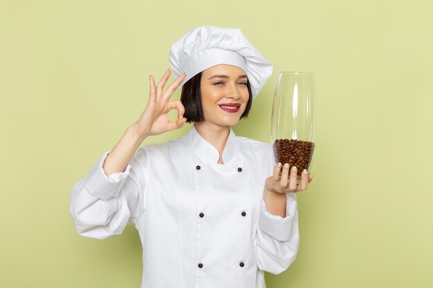 Vista frontal de una joven cocinera en traje de cocinero blanco y gorra sosteniendo un frasco con semillas de café en la pared verde trabajo de dama cocina color