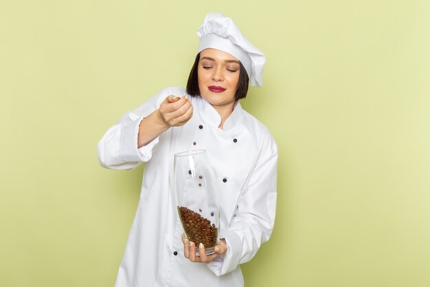 Vista frontal de una joven cocinera en traje de cocinero blanco y gorra sosteniendo un frasco con semillas de café en la pared verde trabajo de dama cocina color