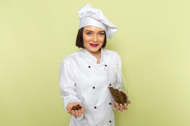 Una vista frontal joven cocinera en traje de cocinero blanco y gorra sosteniendo un frasco con semillas de café sonriendo en la pared verde trabajo de dama cocina color