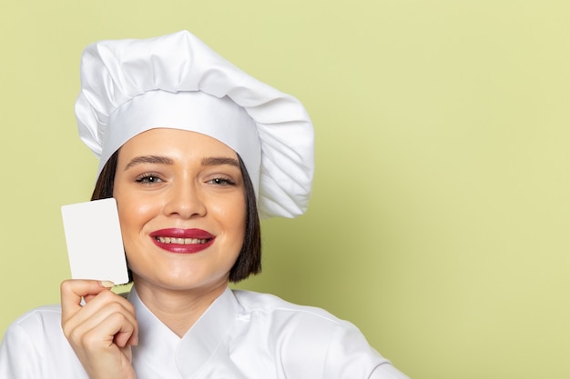Una vista frontal joven cocinera en traje de cocinero blanco y gorra sosteniendo una tarjeta blanca con una sonrisa en la pared verde trabajo de dama cocina color