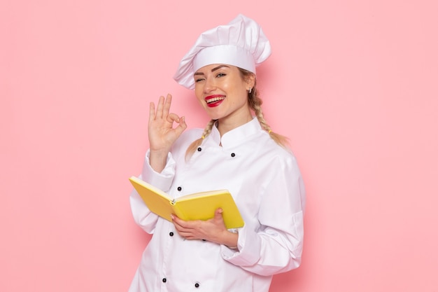 Vista frontal joven cocinera en traje de cocinero blanco leyendo cuaderno amarillo con una sonrisa en el cocinero espacial rosa