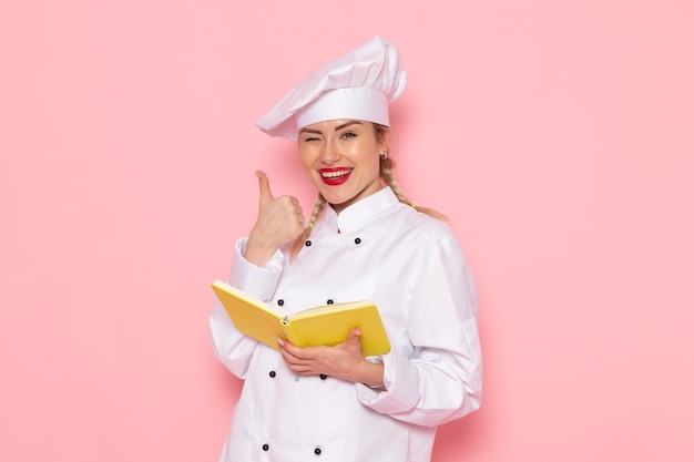 Foto gratuita vista frontal joven cocinera en traje de cocinero blanco leyendo cuaderno con sonrisa en el cocinero espacial rosa