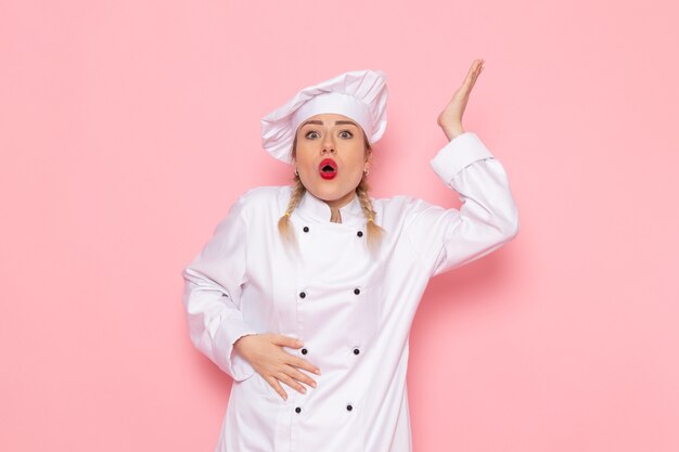 Vista frontal joven cocinera en traje de cocinero blanco posando con expresión de sorpresa en el espacio rosa cocina trabajo trabajo de cocina
