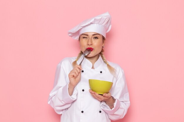 Vista frontal joven cocinera en traje de cocinero blanco posando sonriendo y sosteniendo un plato verde probándolo en el espacio rosa cocina trabajo trabajo cocina