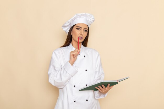 Vista frontal de la joven cocinera en traje de cocinero blanco sosteniendo el cuaderno y pensando en la pared blanca clara