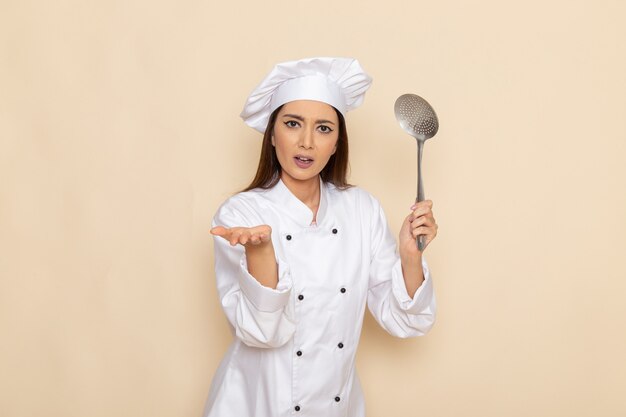 Vista frontal de la joven cocinera en traje de cocinero blanco sosteniendo una cuchara grande de plata en la pared blanca clara