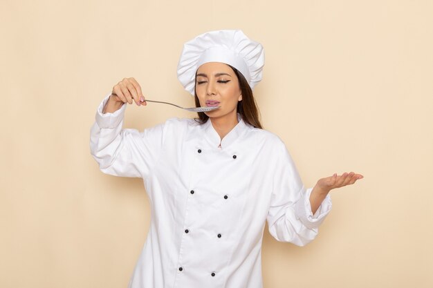 Vista frontal de la joven cocinera en traje de cocinero blanco sosteniendo una cuchara de plata grande degustando en la pared blanca