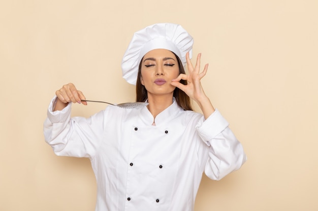 Vista frontal de la joven cocinera en traje de cocinero blanco sosteniendo una cuchara de plata grande en la pared blanca