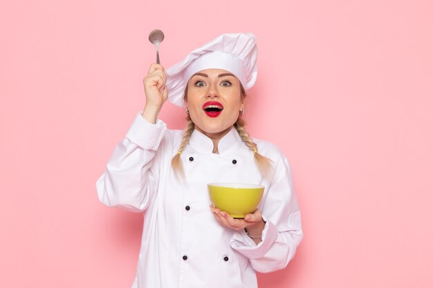 Vista frontal joven cocinera en traje de cocinero blanco sosteniendo una cuchara y un plato verde en el cocinero espacial rosa