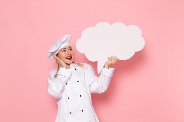 Vista frontal joven cocinera en traje de cocinero blanco sosteniendo un enorme cartel blanco con deleite en el espacio rosa cocinero