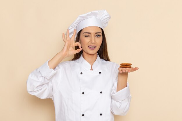 Vista frontal de la joven cocinera en traje de cocinero blanco sosteniendo galletas y guiñando un ojo en la pared blanca clara