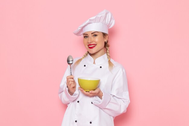 Vista frontal joven cocinera en traje de cocinero blanco sosteniendo placa verde con sonrisa en el cocinero espacial rosa