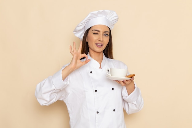 Vista frontal de la joven cocinera en traje de cocinero blanco sosteniendo una taza de café y guiñando un ojo en la pared blanca