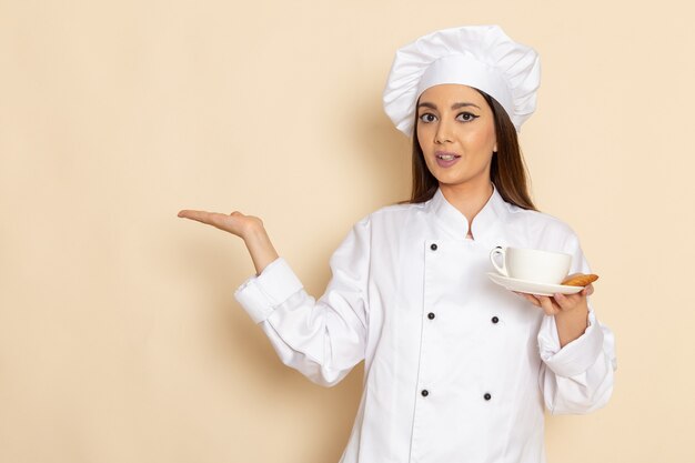 Vista frontal de la joven cocinera en traje de cocinero blanco sosteniendo una taza de café en la pared blanca clara