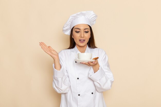 Vista frontal de la joven cocinera en traje de cocinero blanco sosteniendo una taza de café en la pared blanca clara