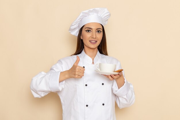 Vista frontal de la joven cocinera en traje de cocinero blanco sosteniendo una taza de café y sonriendo en la pared blanca