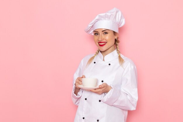 Vista frontal joven cocinera en traje de cocinero blanco sosteniendo una taza de café con una sonrisa en el cocinero espacial rosa