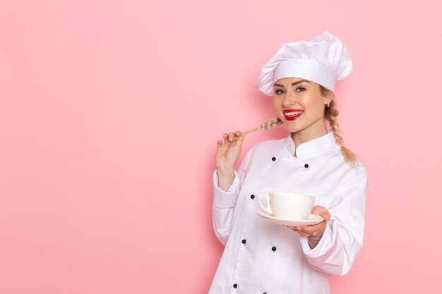 Vista frontal joven cocinera en traje de cocinero blanco sosteniendo una taza con té en el espacio rosa cocinero