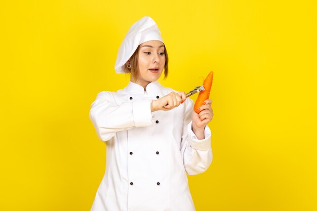 Una vista frontal joven cocinera en traje de cocinero blanco y tapa blanca sosteniendo y limpiando carrott naranja sobre el amarillo