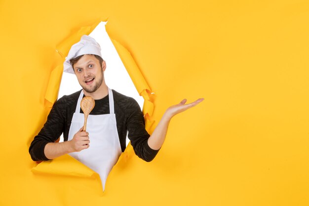 Vista frontal joven cocinero en capa blanca sosteniendo una cuchara de madera sobre el fondo amarillo cocina de color foto trabajo de cocina comida de hombre blanco